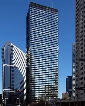 Ground view of a black building's two rectangular, reflective glass facades set in front of a dark blue sky and surrounded by other tall buildings; the smaller facade is bisected by black, inset, crisscrossed beams