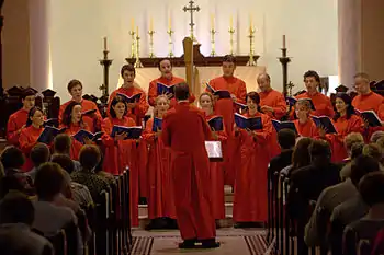 Foto de un coro con túnicas rojas en el presbiterio con el altar detrás de ellos. Sobre el altar velas y cruz de bronce y el maestro de coro volteando a los cantantes dando la espalda a la cámara.
