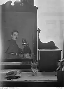 Una cómoda con un espejo grande y una botella de vino, un vaso y unos papeles que se sientan en la parte superior. Un hombre con uniforme militar se puede ver en el reflejo del espejo. Él tiene su mano en el bolsillo, y está utilizando una cámara en un trípode.