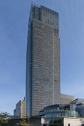 Ground-level view of a tall, mostly glass facade set in front of a blue, cloudy sky; a smaller, circular building is in the foreground