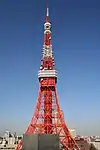 The orange and white lattice frame of Tokyo Tower rises up in front of a clear, blue sky.
