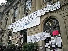 Fotografía del frontis de la Casa Central de la Universidad Católica de Chile cubierto con lienzos y pancartas que cuelgan de la reja de la puerta principal y del segundo piso. Uno de esos lienzos dice "Toma feminista"..