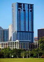 A view from Zilker Park of the building; the tower is topped out; the building is rectangular, and there are no setbacks; the building has a reflective blue facade; the tower appears to look like a tall rectangle stacked on top of another smaller rectangle.