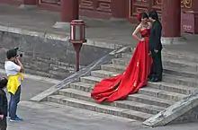 A woman wearing a long, ornate red dress stands next to a man in a black suit on some short stone steps while another man photographs them from the foot of the stairs