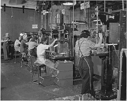Una foto de la época de la II Guerra Mundial de cinco mujeres trabajando en taladros, todas vestidas con pantalones, camisas de trabajo, sobreros y mandiles.