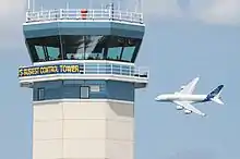 Nueva torre de control del Aeropuerto de Oshkosh con el texto "la torre de control más ocupada del mundo". Al fondo sobrevuela un Airbus A380.