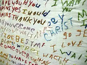 A white cloth with seemingly random, unconnected text sewn into it using multiple colors of thread