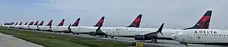 Many in line airplanes with the Delta Air Lines logo on the tail, parked on pavement behind a fence.