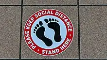 Circular sign with a silhouette of bare feet, with text in a red border that says "Please keep social distance • Stand here"