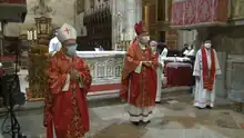 Two men in masks, wearing mitres and red vestments, stand in front of an altar. Altar servers, deacons, and priests in the background similarly wear masks.