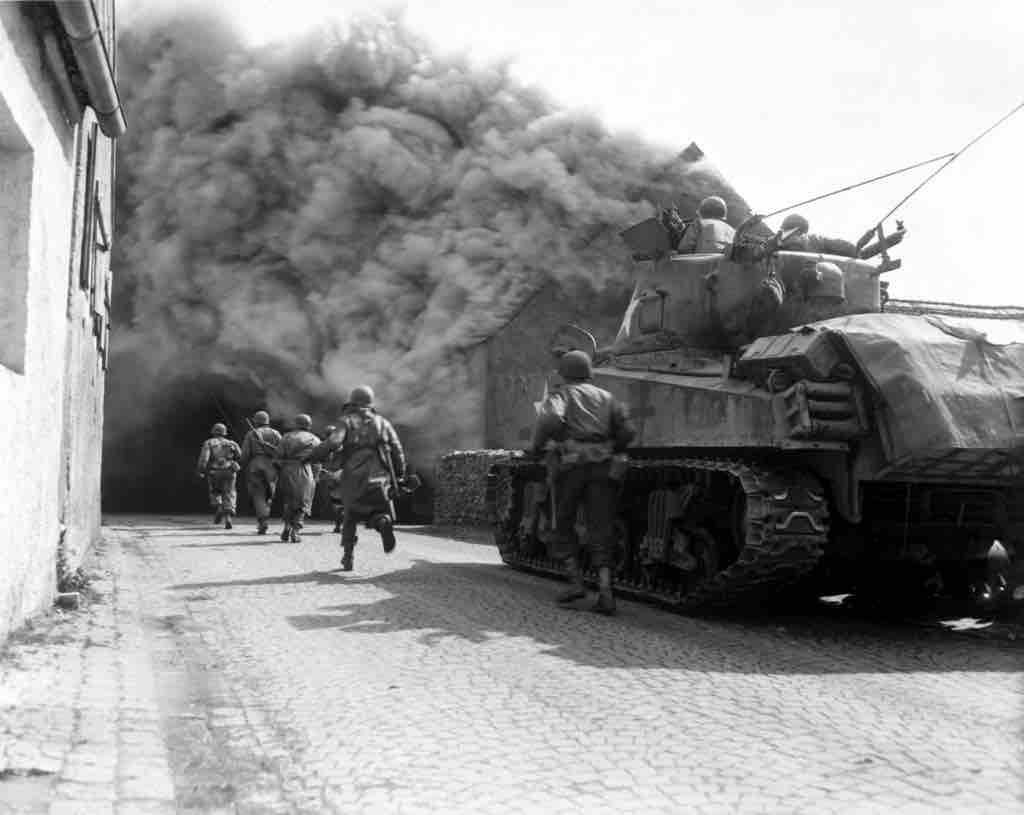 
United States Army soldiers supported by a M4 Sherman tank move through a smoke filled street in Wernberg, Germany during April 1945.
