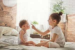 Older sister sitting on a bed face-to-face with baby sister, lovingly touching her nose