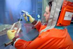 Man is wearing a powered air purifying respirator, gloves, and solid-front gown. He is working within a BSC, and self-closing, locked doors are in the background.