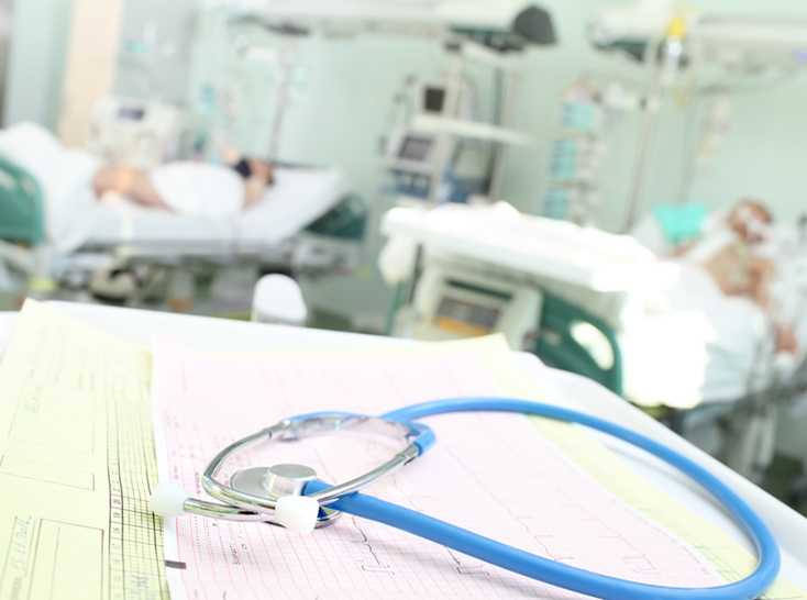 A blue and silver stethoscope is resting on top of white papers. In the background, patients rest in hospital beds